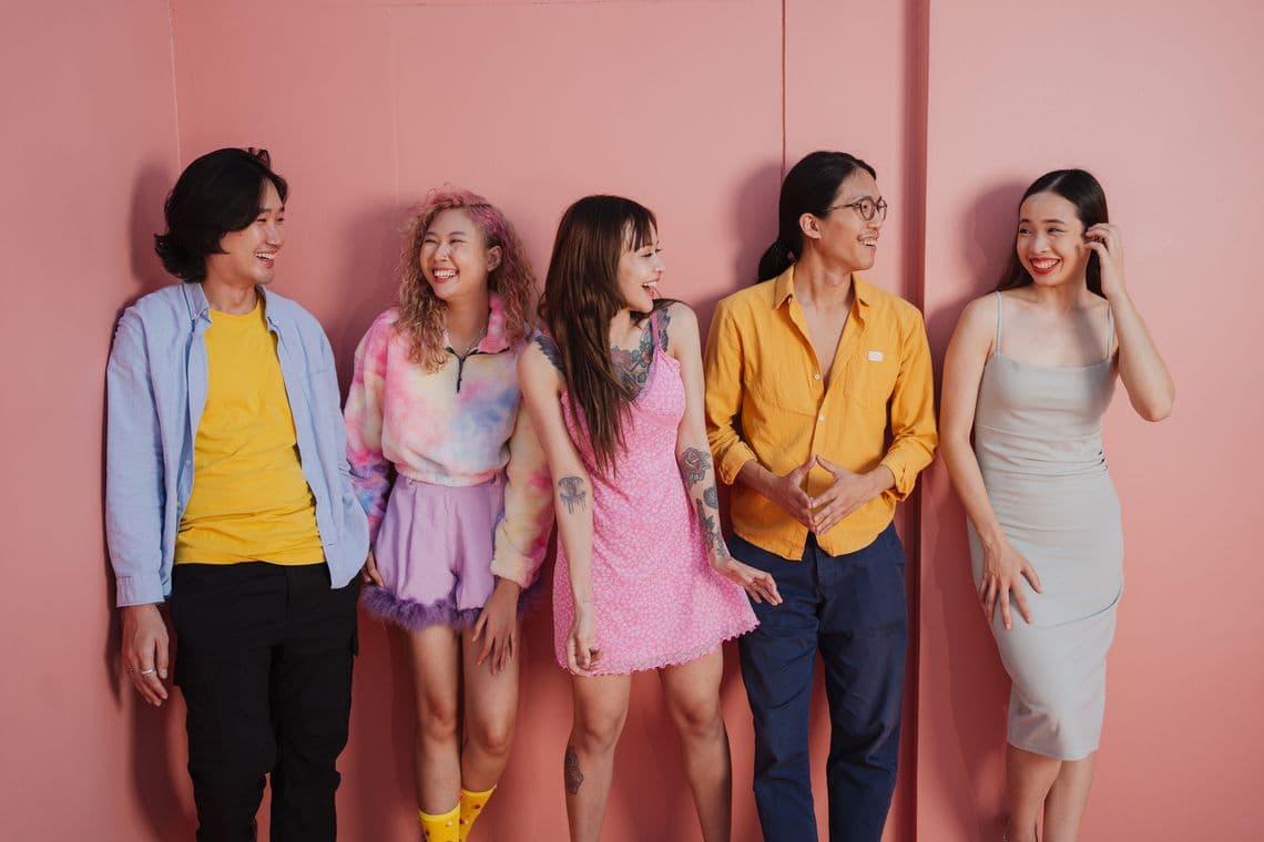 Five people smiling and chatting in front of a pink wall, dressed in colorful and casual outfits.