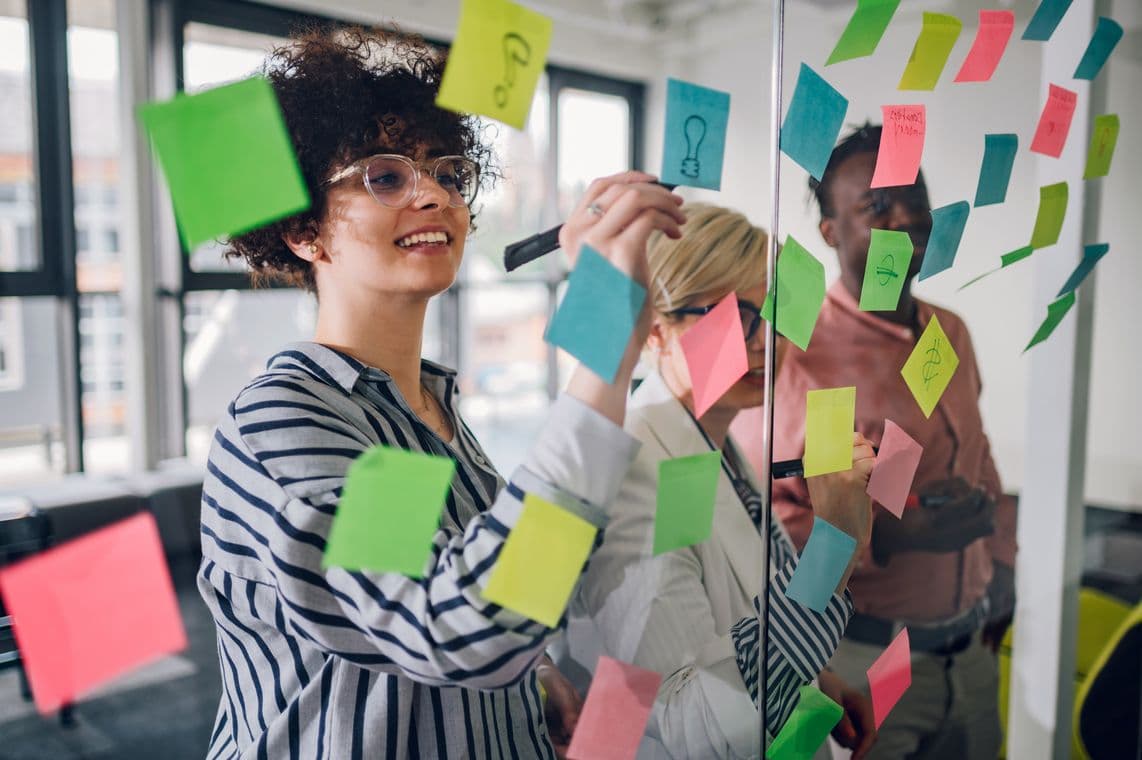 People brainstorming in an office, writing on colorful sticky notes on a glass wall. Bright, collaborative atmosphere.