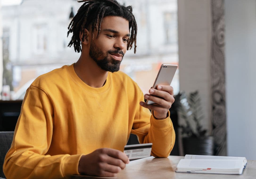 Man in yellow sweater holding a credit card and smartphone, smiling, seated at a table with an open notebook.