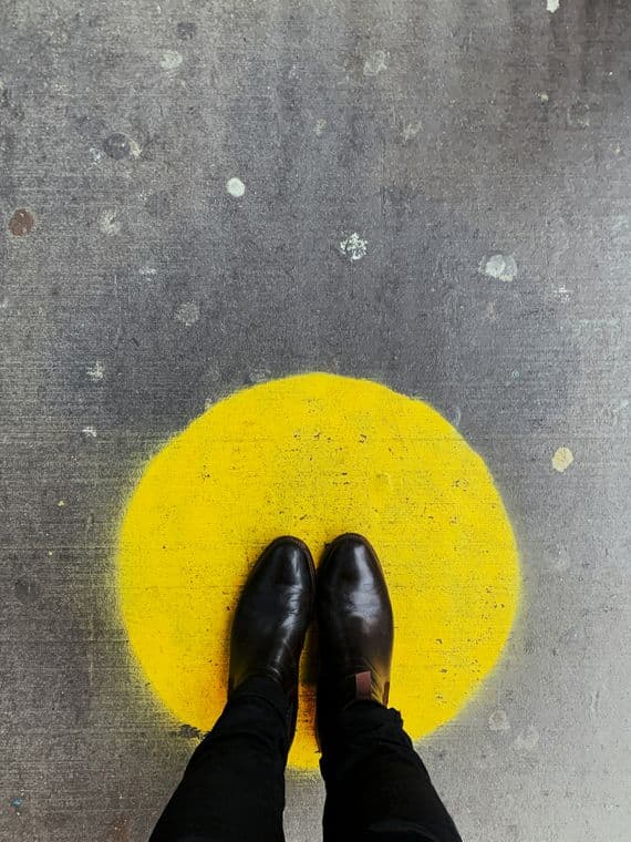 Person wearing black shoes standing on a yellow circle painted on a gray, speckled concrete floor.