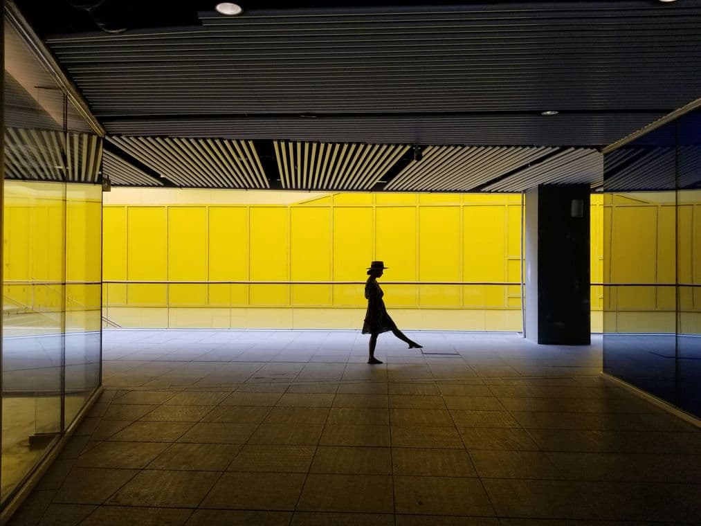 Silhouette of a person walking in a corridor with bright yellow walls and a striped ceiling, casting a shadow on the tiled floor.