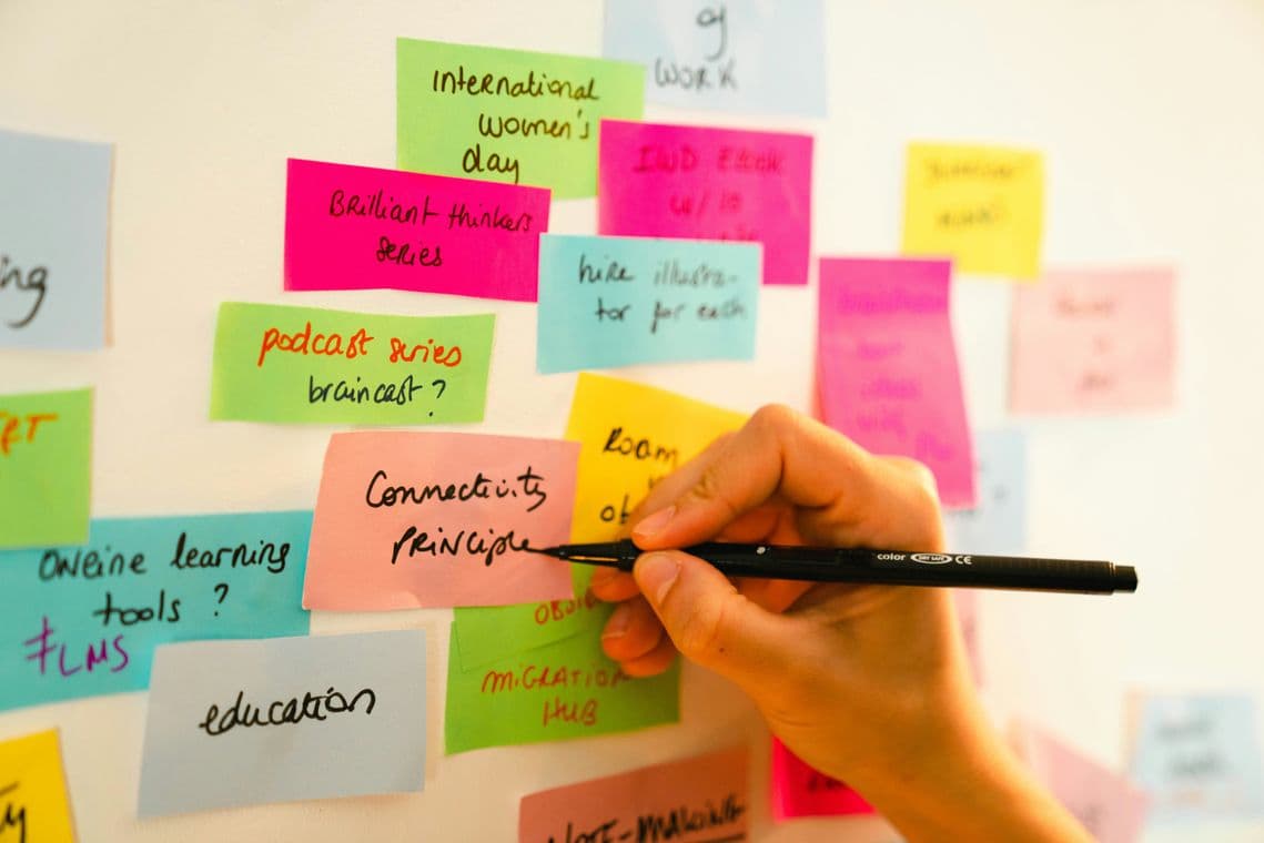 A hand writes on a pink sticky note among various colorful notes on a whiteboard, featuring phrases like "Connectivity Principles" and "education."